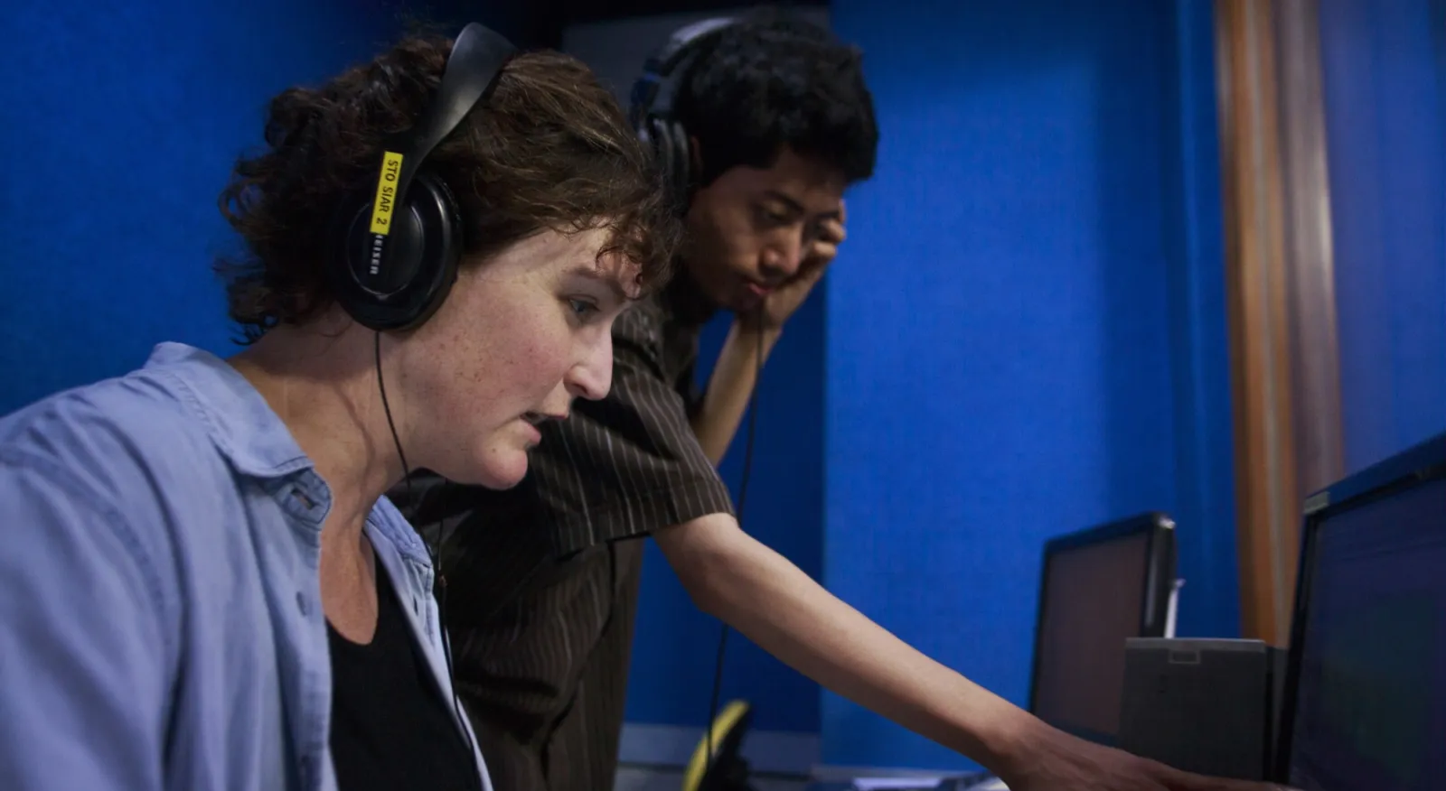 Two people work together in a audio studio, both wear headphones and look at a soundboard