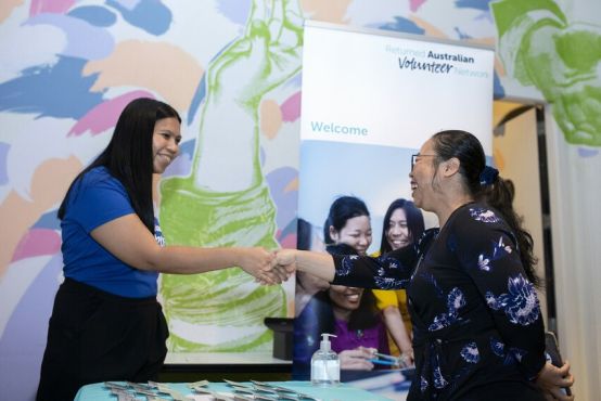 A lady with long dark hair who is wearing a program t-shirt shakes hands with a woman in a dark blue top. They stand in front of an Alumni banner