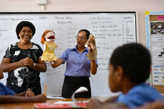Two women stand in front of a classroom, in front of a white board. They are both smiling and engaged with two hand puppets, one of a redheaded girl and the other a kangaroo.