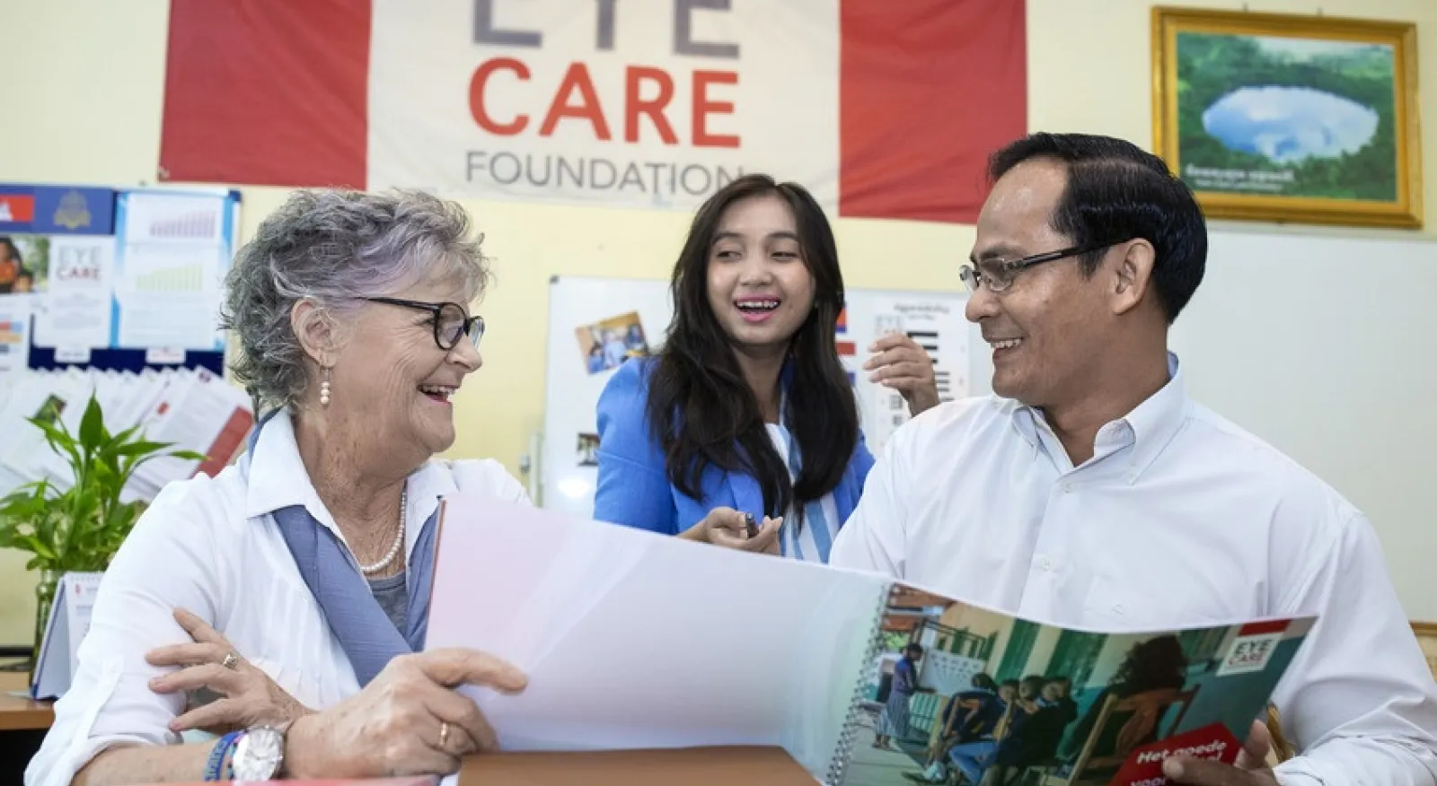 Three people looking at a brochure and smiling. A sign behind them all says 'Eye Care Foundation'
