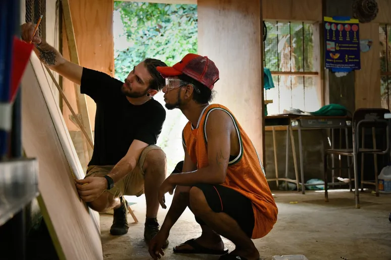 Two people crouched down measuring a piece of timber in a workshop.