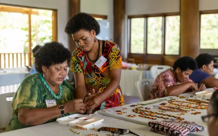 Two people working on beading together. One woman sits at the table, and the other stands next to her.