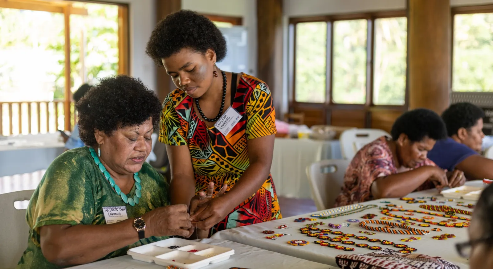 Two people working on beading together. One woman sits at the table, and the other stands next to her.