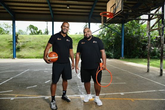 Two people standing side by side in front of a basketball court. One man has a basketball in his hand, the other has a hoop in his hand.