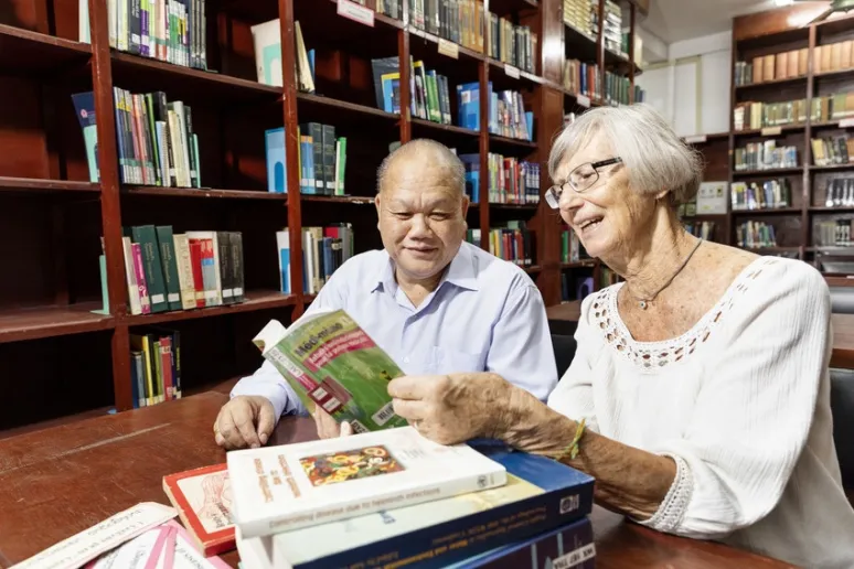 Two people sit in a library together, looking at a book on a desk. Shelves behind them host an array of books.