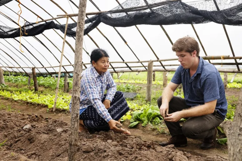 A person in a checked shirt and traditional skirt squats in a greenhouse with a person in a short-sleeved blue shirt, they both examine the soil