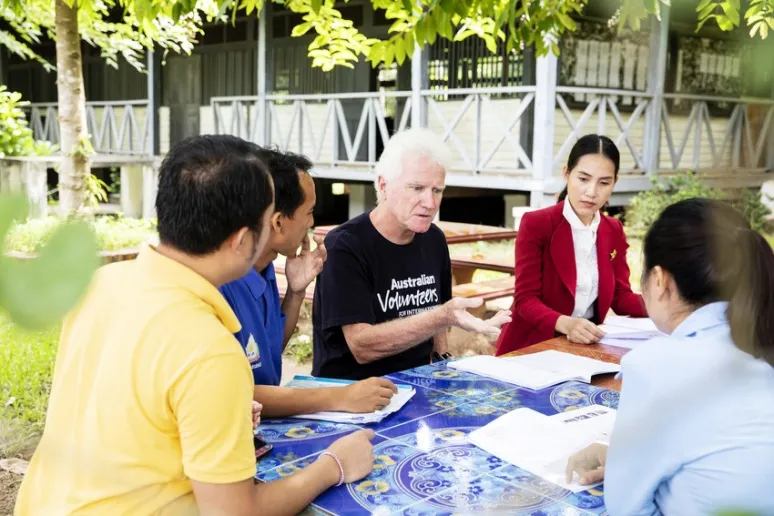 A man with white hair in an Australian volunteers t-shirt sits at a table with two other men and two women, they all look at workbooks in front of them.