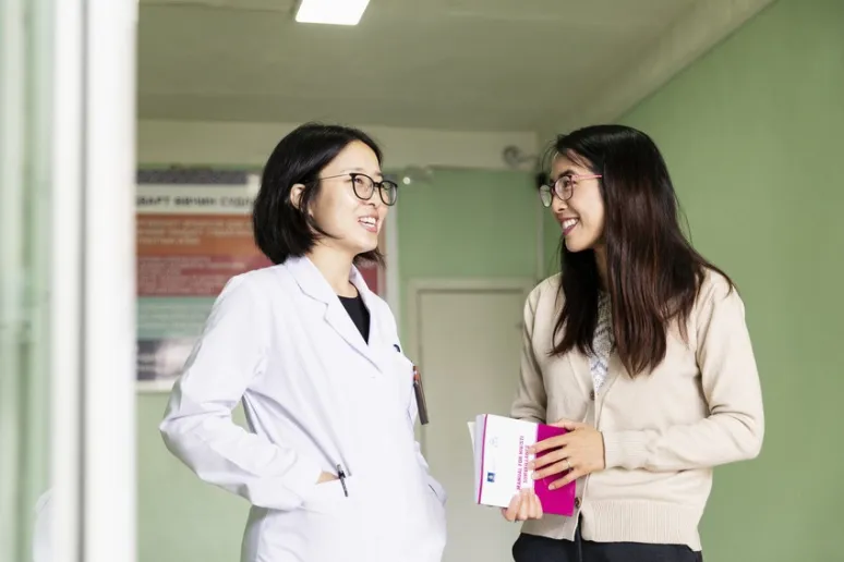 Two women stand in a room talking. One of them is wearing a white lab coat.