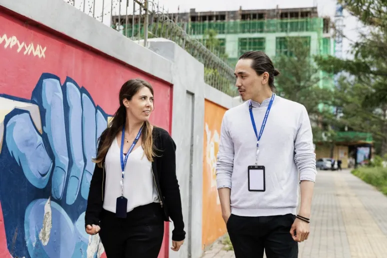 Two people walk down a street, past a mural on the wall