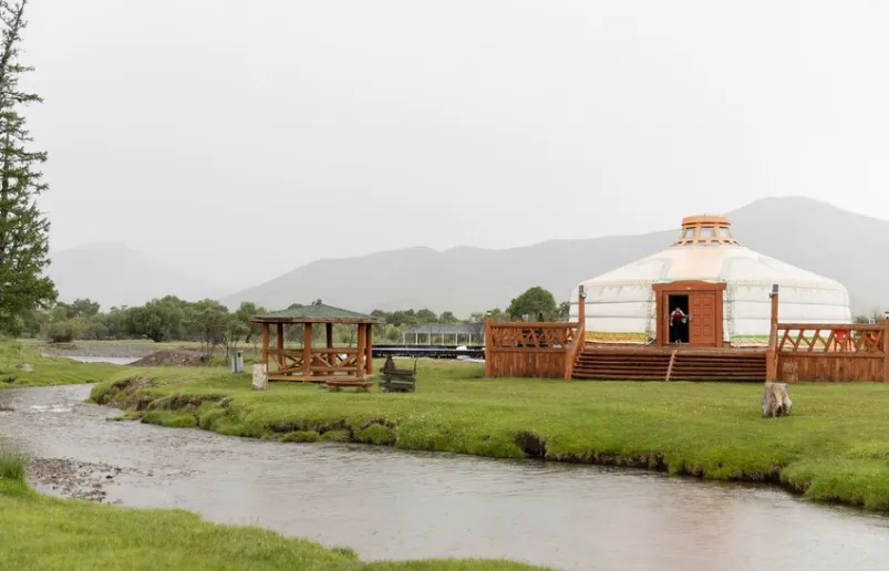 A Mongolian landscape with a river in the foreground and mountains in the background. It shows a yurt, a traditional Mongolian round dwelling.