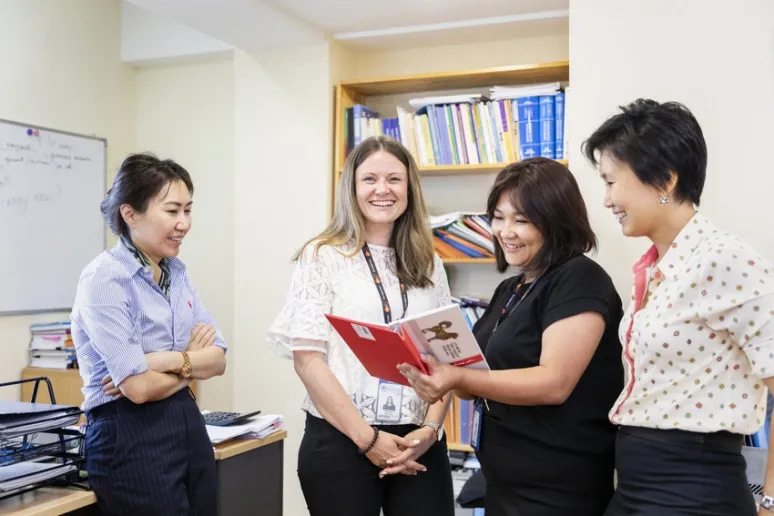 Four people stand in an office. Three look at a book, and one looks at the camera