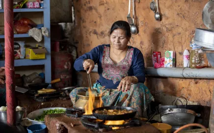 A woman sits in a kitchen cooking food with flames around her pots. She is wearing a traditional Nepalese dress in blue with embroidery around the neck.