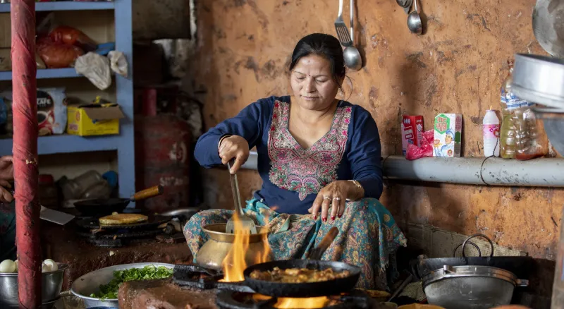 A woman sits in a kitchen cooking food with flames around her pots. She is wearing a traditional Nepalese dress in blue with embroidery around the neck.