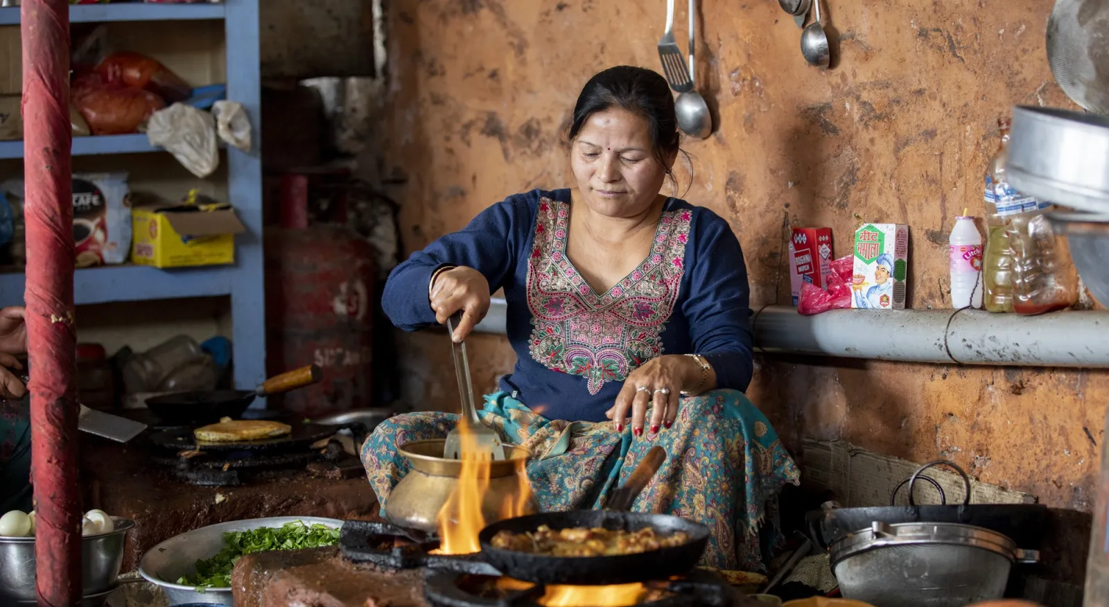 A woman sits in a kitchen cooking food with flames around her pots. She is wearing a traditional Nepalese dress in blue with embroidery around the neck.