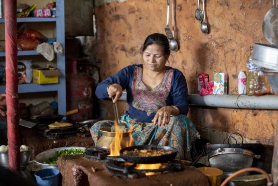 A woman sits in a kitchen cooking food with flames around her pots. She is wearing a traditional Nepalese dress in blue with embroidery around the neck.