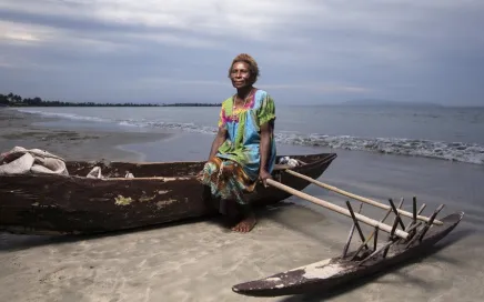 A lady in a brightly coloured dress sits on an outrigger canoe on a beach with water behind her