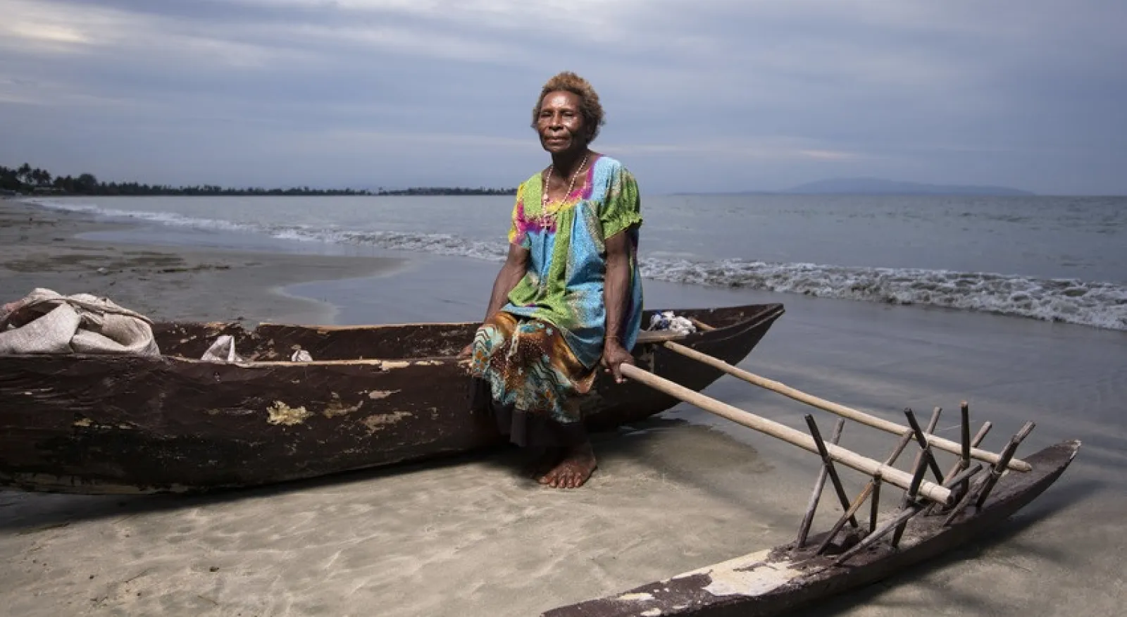 A lady in a brightly coloured dress sits on an outrigger canoe on a beach with water behind her