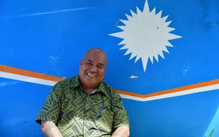 A man in a patterned green shirt sits smiling in front of a painted Republic of Marshall Island flag. It is light blue with a white star-like shape, with a slim orange and white stripe.