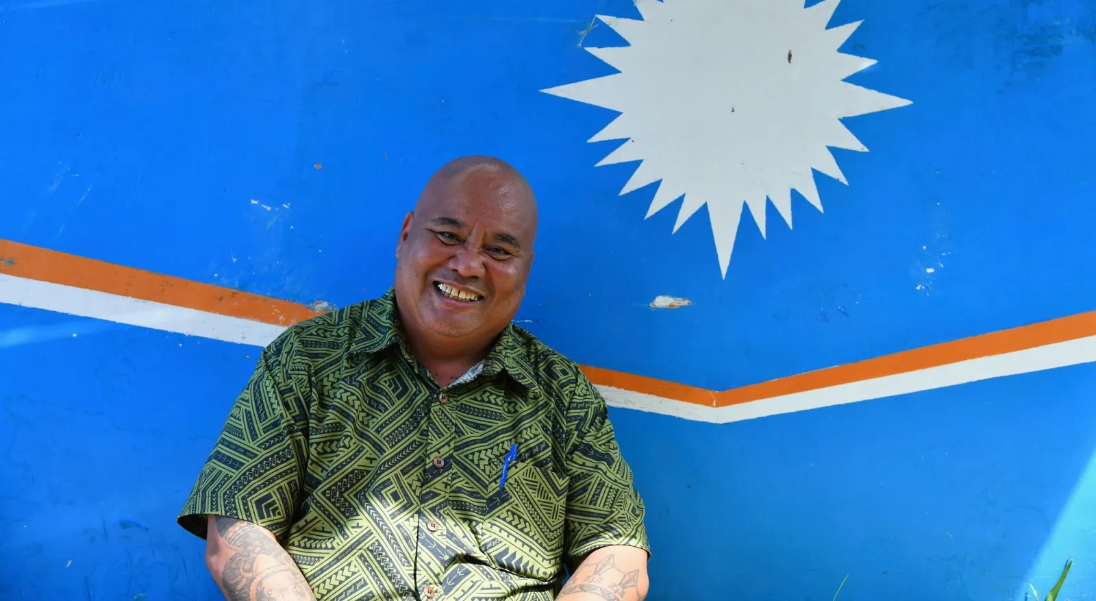 A man in a patterned green shirt sits smiling in front of a painted Republic of Marshall Island flag. It is light blue with a white star-like shape, with a slim orange and white stripe.