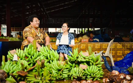 Two people sit behind a large pile of bananas at a marketplace. They are in traditional Samoan clothing, one wears a bright yellow print and the other a light blue.