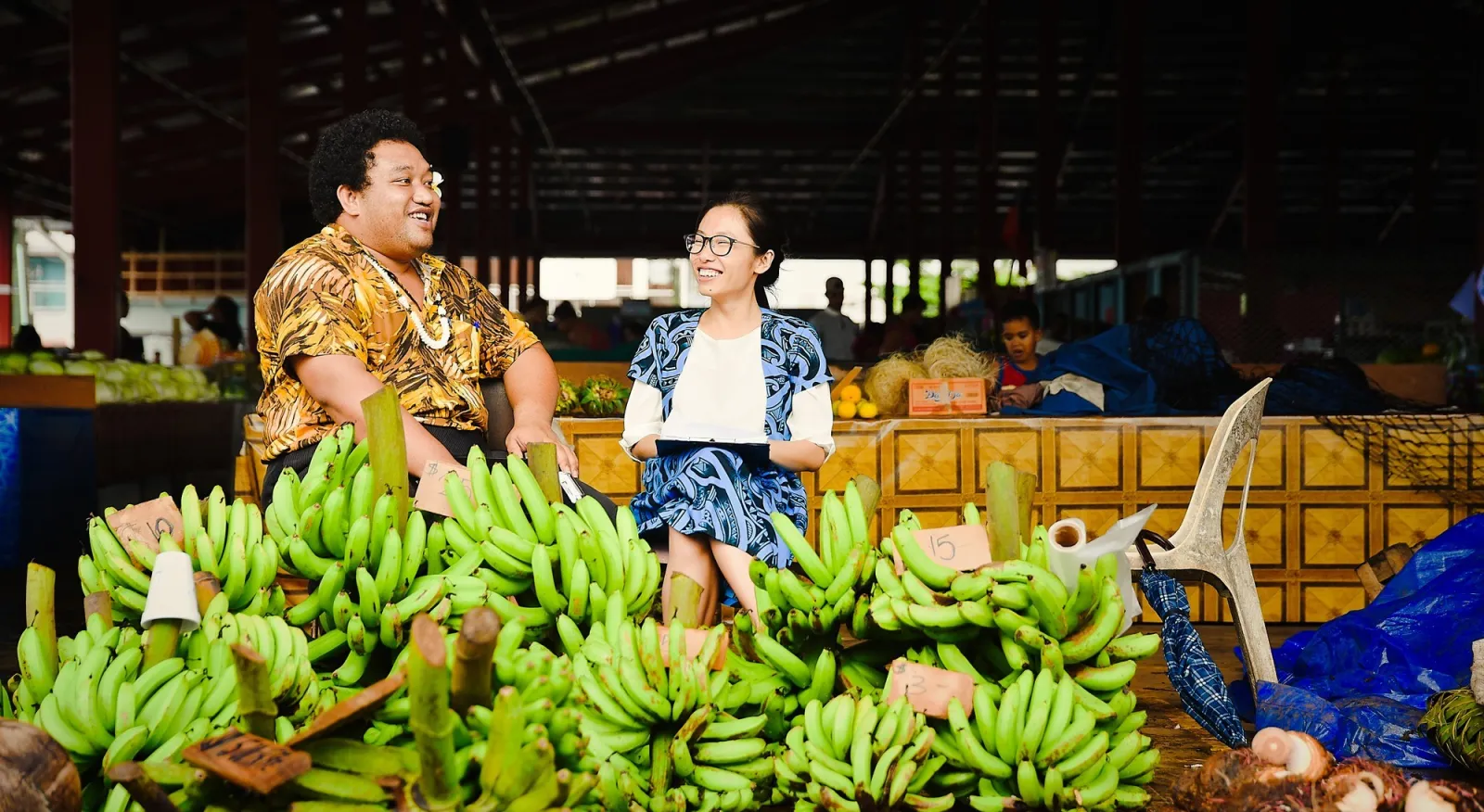 Two people sit behind a large pile of bananas at a marketplace. They are in traditional Samoan clothing, one wears a bright yellow print and the other a light blue.