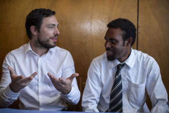 Two people sit in button up shirts against a brown wall. They are smiling and talking, one is holding his hands up mid-expression.