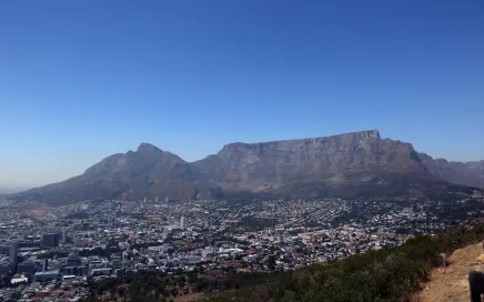 A landscape photo of Table Mountain in South Africa. The mountain has a large cityscape below it, and a bright blue sky above. The rock itself is a dark brown/red.