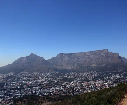 A landscape photo of Table Mountain in South Africa. The mountain has a large cityscape below it, and a bright blue sky above. The rock itself is a dark brown/red.
