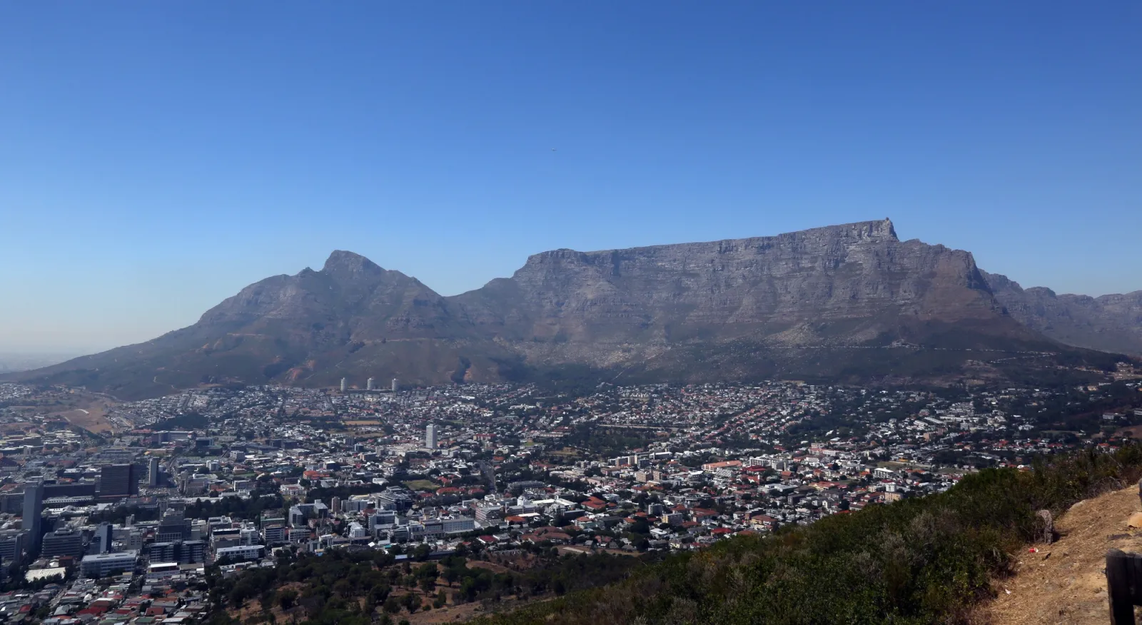 A landscape photo of Table Mountain in South Africa. The mountain has a large cityscape below it, and a bright blue sky above. The rock itself is a dark brown/red.