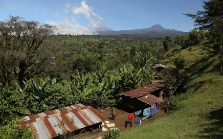 A series of buildings are below a ridge, behind them lays a long green landscape, with Kilimanjaro rising in the distance.