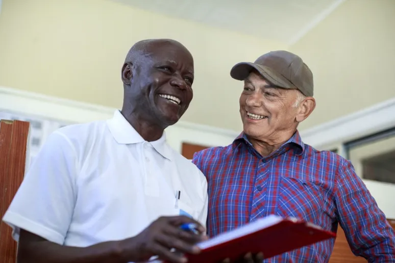 A Black man and a white man look at each other smiling. The black man is holding a red clipboard.