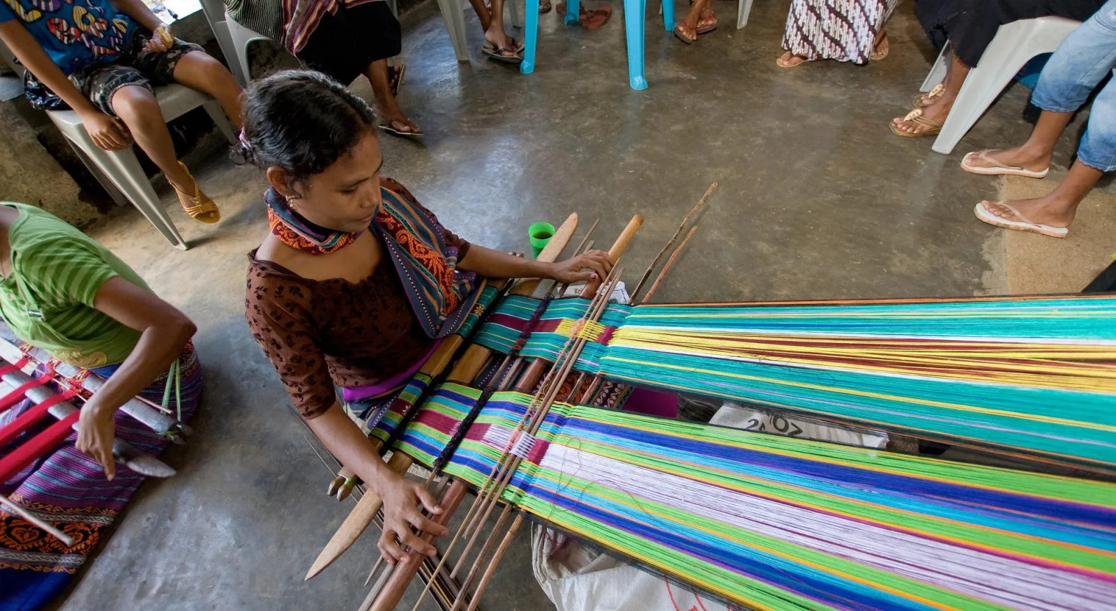 A woman skillfully creating vibrant fabric using a traditional weaving machine. You can see other people sitting further back in the photo, watching her work.
