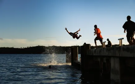 A group of people joyfully leaping into the water from a pier in Tonga, creating a big splash. One man is mid-jump above the water.