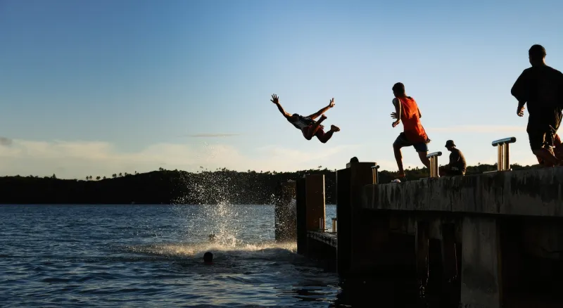 A group of people joyfully leaping into the water from a pier in Tonga, creating a big splash. One man is mid-jump above the water.