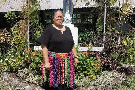 A lady stands in front of a garden, she is wearing a brightly coloured woven skirt