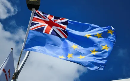 The flag of Tuvalu blows in the breeze against a blue sky. The flag is baby blue, with 9 gold stars across the body and the Union Jack in the upper right hand corner.