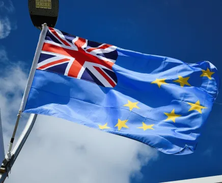 The flag of Tuvalu blows in the breeze against a blue sky. The flag is baby blue, with 9 gold stars across the body and the Union Jack in the upper right hand corner.