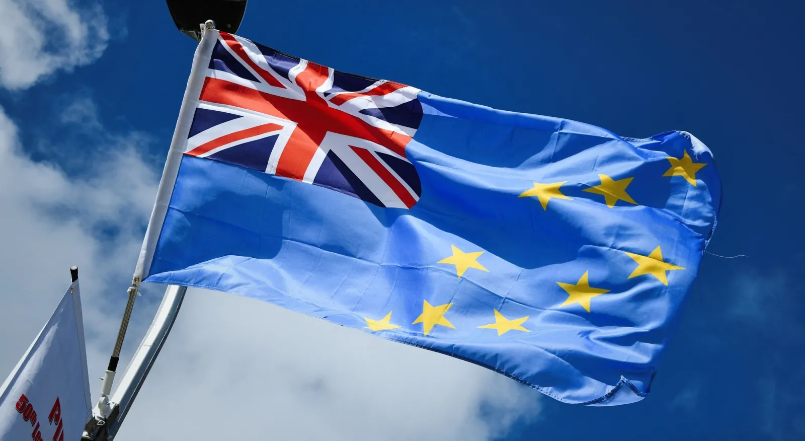 The flag of Tuvalu blows in the breeze against a blue sky. The flag is baby blue, with 9 gold stars across the body and the Union Jack in the upper right hand corner.