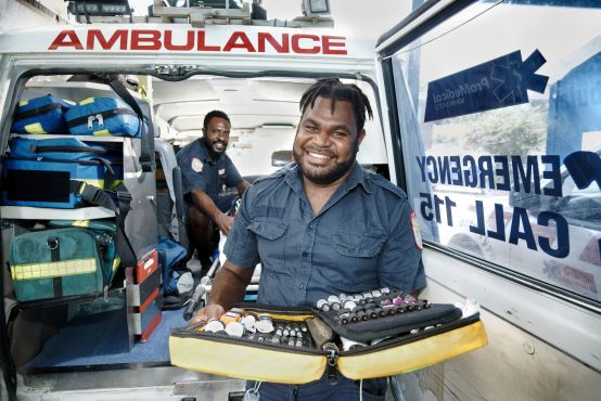 One man stands in the open door of an ambulance, and one man sits inside the ambulance. Both wear medical uniforms.