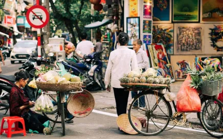 A busy streetscape in Vietnam shows two people with bicycles carrying fruit.