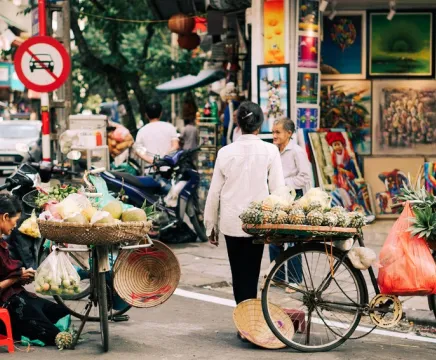 A busy streetscape in Vietnam shows two people with bicycles carrying fruit.