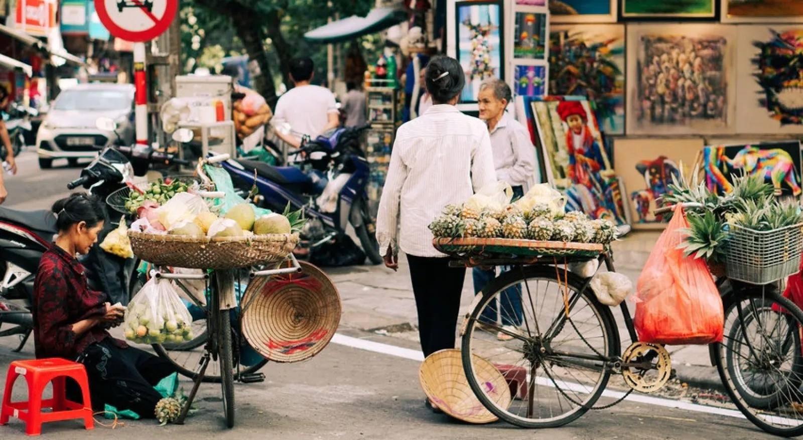 A busy streetscape in Vietnam shows two people with bicycles carrying fruit.