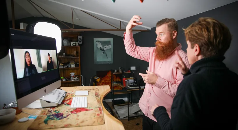 A man and a woman are signing in ASL to two women on a computer screen on Zoom. The man has red hair and a thick red beard and the camera can see his face. The woman is facing away from the camera, looking at her colleague.
