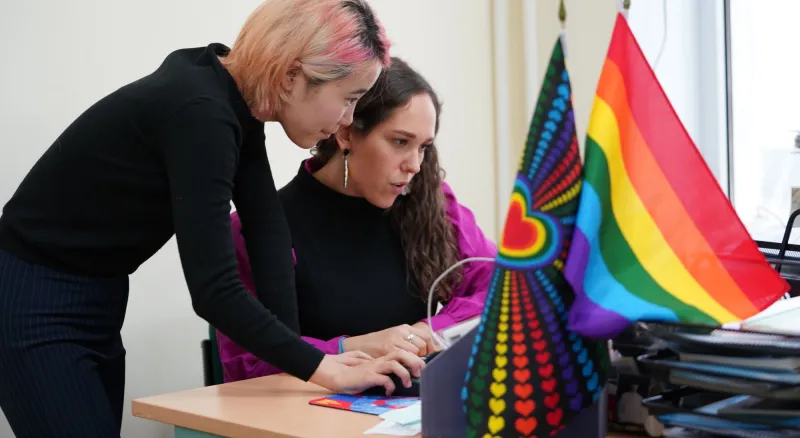 Two women sit at a desk working and smiling. There are two LGBTIQ+ pride flags on the desk.
