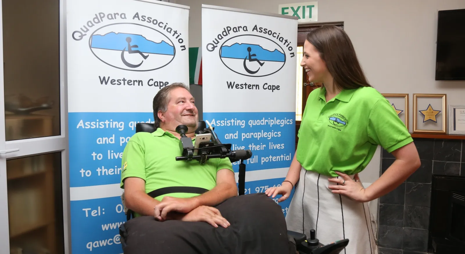 A man and a woman are talking and smiling in front of a QuadPara Association sign. They are both wearing bright green shirts and the man is in a wheelchair