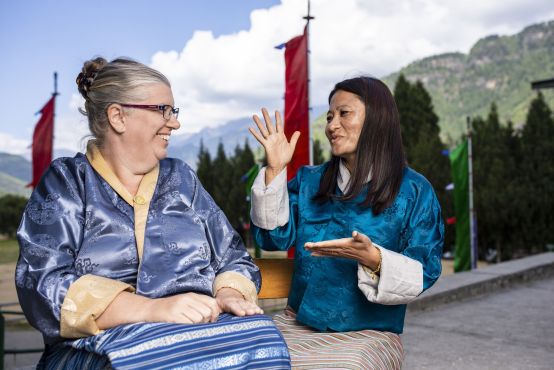 Two ladies in traditional Bhutanese dress face each other, one uses sign language. They are outside and behind them are mountains and flags