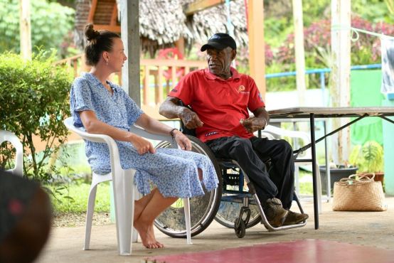 A lady in a blue dress sits on a white chair, she talks to a man in a red top who is in a wheelchair. They sit outside on a balcony, behind them are plants and a thatched hut