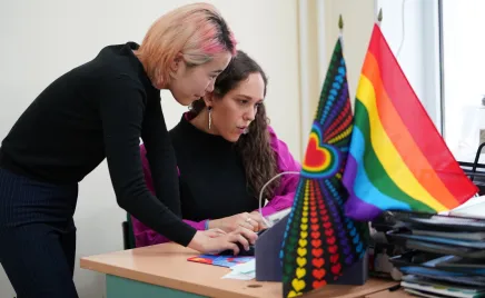 Two women sit at a desk working and smiling. There are two LGBTIQ+ pride flags on the desk.