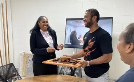 A man in an Australian Volunteers Program Indigenous Pathways t-shirt hands a gift to a lady wearing a suit. In the background is the Indigenous Australian flag