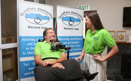 A man and a woman are talking and smiling in front of a QuadPara Association sign. They are both wearing bright green shirts and the man is in a wheelchair